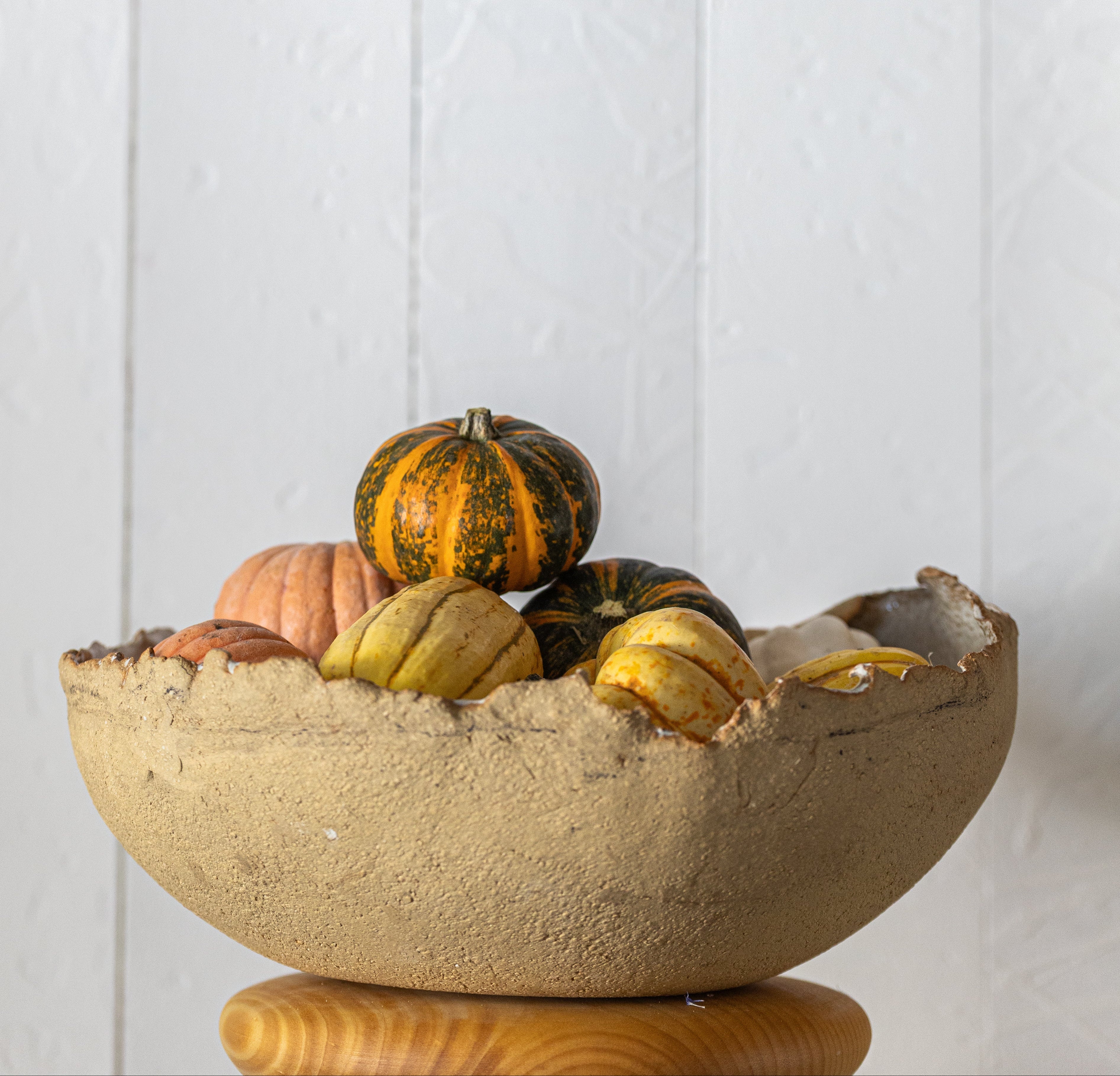 A rustic beige ceramic bowl filled with assorted gourds and small pumpkins, displayed on a wooden stand against a white paneled wall.